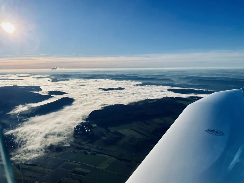 Wolkenmeer über Tälern aus dem Cockpit gesehen, Tragfläche im Vordergrund